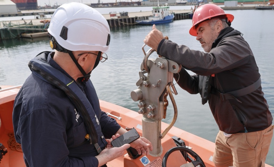 Two CRALOG inspectors on a lifeboat checking the hook release, one recording the result on the CRALOG app